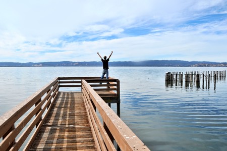 A man standing at the edge of a long narrow wooden pier above the calm ocean water with outstretched victorious arms.の写真素材