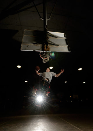 Basket and basektball player jumping with ball and aiming at basket with liights turned on at night