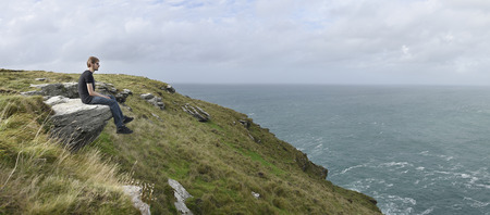 Man sitting on a rock on a grass hill cliff overlooking the sea at Tintagel Castle, Cornwall, UKの写真素材