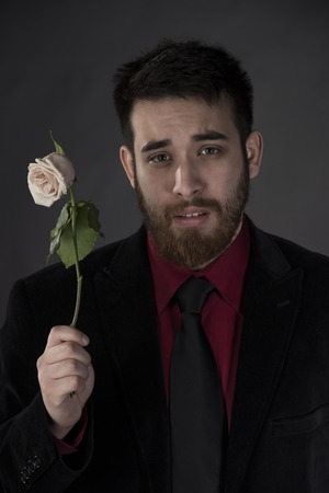 Close up Sad Young Man in Elegant Formal Wear Holding Withered Rose on One Hand While Looking at the Camera. Isolated on Gray Background.の写真素材