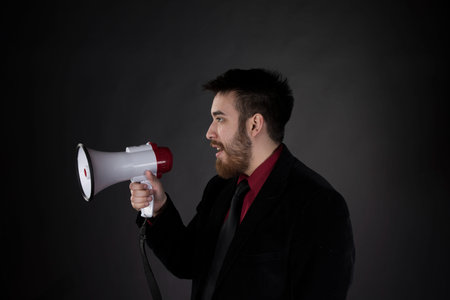 Half Body Shot of Young Goatee Man in Side View, Wearing Formal Wear, Speaking Using Megaphone on Gradient Gray Background.の写真素材