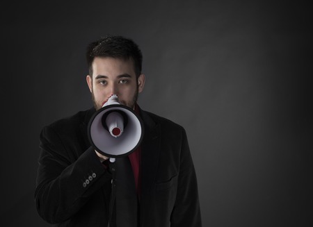 Close up Young Gorgeous Man Wearing Formal Suit Holding Megaphone While Looking at the Camera on Gradient Gray Background.の写真素材