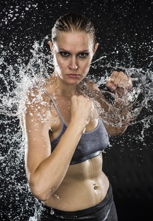 Half Body Shot of an Athletic Woman Doing Combat Pose in Water Splashes, Looking Aggressive at the Camera on a Black Background.の写真素材