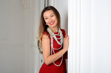 Smiling white woman with long brown and blond hair in white pearl jewelry in long red evening dress on light backgroundの写真素材