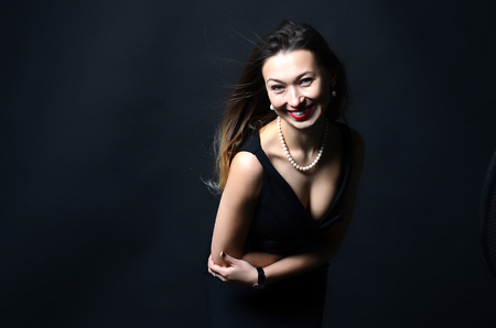 Smiling white woman with long brown and blond hair in white pearl jewelry in long black evening dress on dark backgroundの写真素材