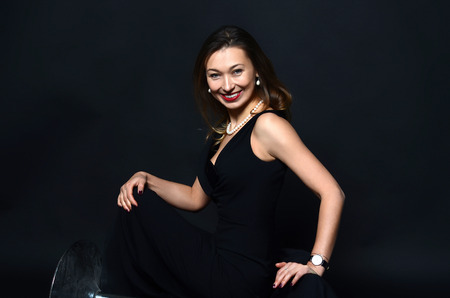 Smiling white woman with long brown and blond hair in white pearl jewelry in long black evening dress on dark backgroundの写真素材