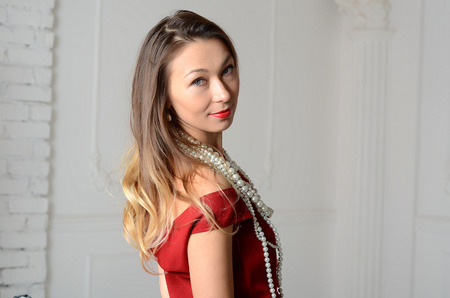 White woman with long brown and blond hair in white pearl jewelry in long red evening dress on light backgroundの写真素材