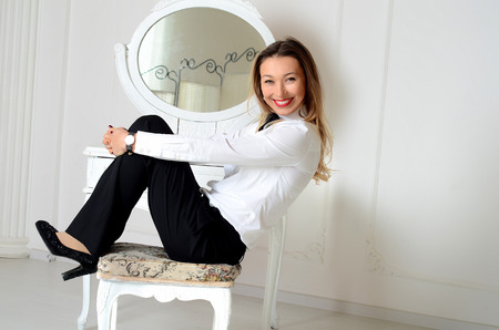 Smiling white woman in white shirt, black pants and black shoes sitting in white studio in front of mirrorの写真素材