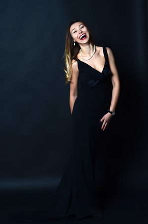 Laughing white woman with long brown and blond hair in white pearl jewelry in long black evening dress on dark backgroundの写真素材