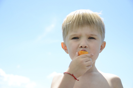 Beautiful white blonde boy with blue eyes eating apricot on the blue sky backgroundの写真素材