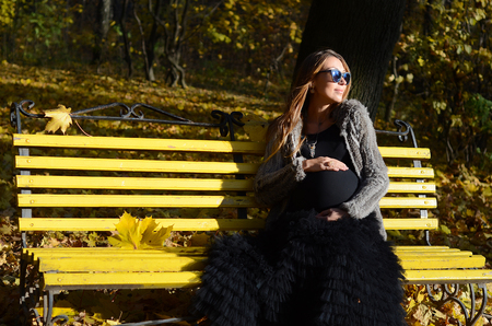 Happy beautiful pregnant white girl with long hair in dark clothing, sunglasses, sitting on the yellow bench in the park, autumn, many yellow leaves, a beautiful sunny dayの写真素材