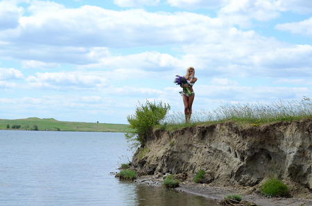 beautiful white blond girl with long hair, blue eyes, wearing a white bikini with a big bouquet of purple flowers, sitting in a field with green grass and flowers on the bank of a lakeの写真素材
