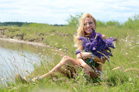 beautiful white blond girl with long hair, blue eyes, wearing a white bikini with a big bouquet of purple flowers, sitting in a field with green grass and flowers on the bank of a lakeの写真素材