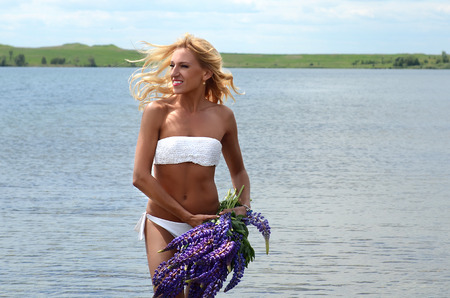 beautiful white blond girl with long hair in white bikini with a big bouquet of purple flowers, standing in the water in a large lakeの写真素材