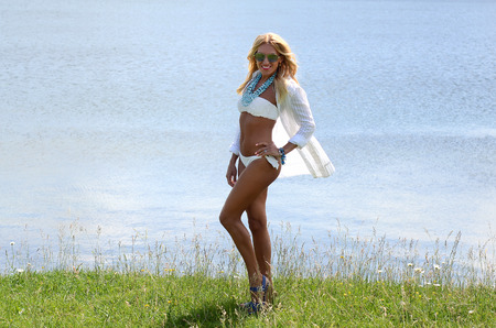 beautiful white blond girl with long hair in a white bikini, a white shirt, sunglasses, blue jewelry, dancing near the lake in the summerの写真素材
