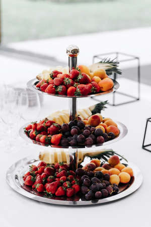 fruit plate. Three floors of fruit on a wedding table with strawberries, pineapple, apricot, grapes on light backgroundの写真素材
