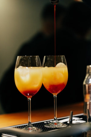 on the bartenders table are two glasses of orange and red cocktails into which the drink is poured....