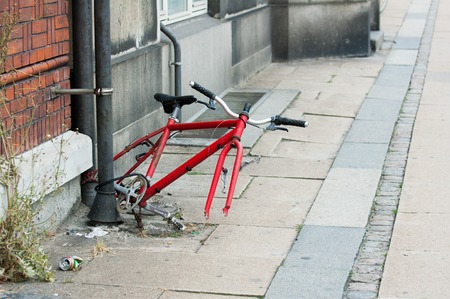 The red frame from the old bicycle with the remnants of the construction elements is tied to a black pipe near the wall of a house on a littered street.の写真素材