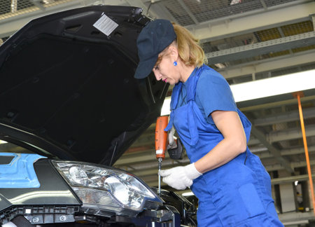 The female collector fixes a detail in podkapotny space of the car. Assembly shop of automobile plantのeditorial素材