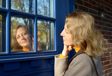 A young woman looks at her reflection on the glass of a blue door