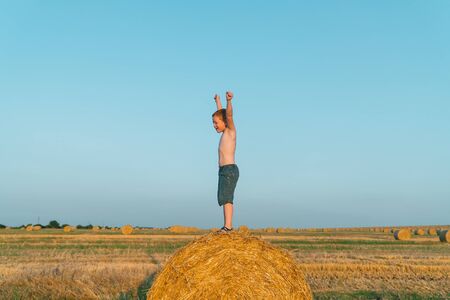 A red-haired boy stands on top of a straw bale on a wheat fieldの写真素材