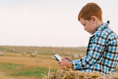 A red-haired boy on top of a straw bale holds a tablet in his hands on a background of hay balesの写真素材