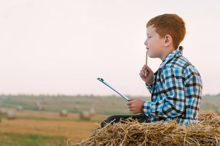 A red-haired boy on top of a straw bale holds a tablet with a letter and a pen in his hands on a background of hay balesの写真素材