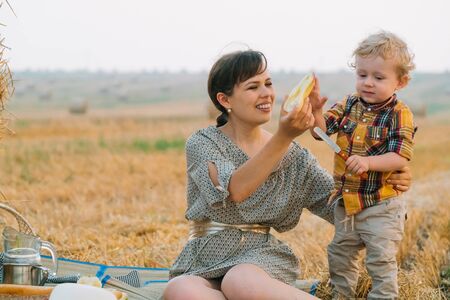 Beautiful young woman with little blond son on picnic near hay bale in the middle of wheat field in summer eveningの写真素材