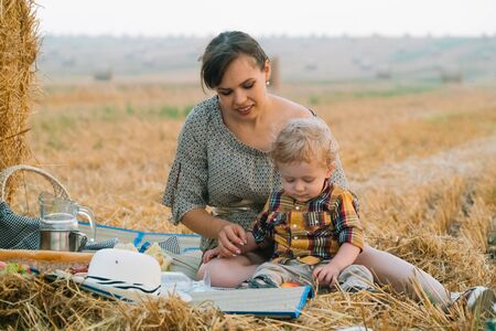 Beautiful young woman with little blond son on picnic near hay bale in the middle of wheat field in summer eveningの写真素材