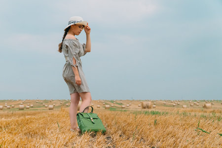 Beautiful young woman in a hat and with a backpack in the middle of a wheat field with bales in a...