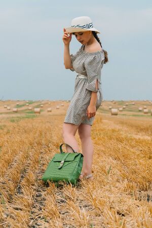 Beautiful young woman in a hat and with a backpack in the middle of a wheat field with bales in a summer eveningの写真素材