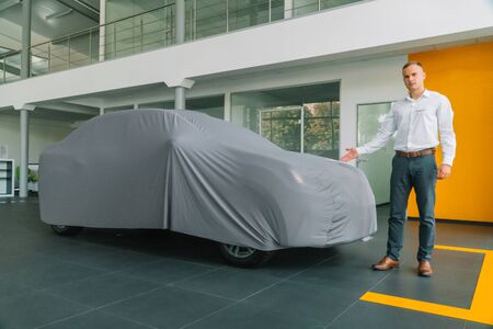 The seller in a car dealership stands near a car covered by a protective coverの写真素材