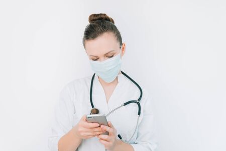 Young nurse in a protective mask uses her smartphone on a white background in a hospitalの写真素材