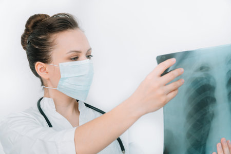 Young caucasian doctor in protective mask examine x-ray of lungs, holding it in hands indoors....