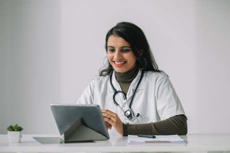 Indian female doctor in a white coat with a stethoscope conducts an online consultation in her officeの写真素材
