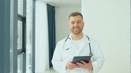 Successful physician in white lab coat with tablet looks at the camera and smiles in hospital officeの写真素材