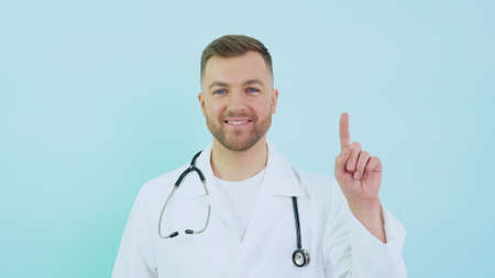Doctor with stethoscope in white coat raises his index finger up to face level on a blue backgroundの写真素材