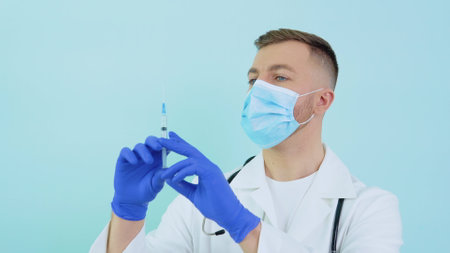 Male doctor in a protective mask with a vaccine syringe in his hands on a blue backgroundの写真素材