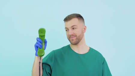A friendly doctor in a green uniform and blue gloves holds an old phone in his hands and points to itの写真素材
