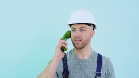 Serviceman in overalls and helmet speaks on an old telephone and consults a client on a blue backgroundの写真素材