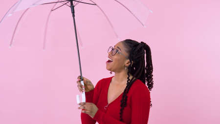 Young african american girl having fun with transparent umbrella in photo studio. Teen girl in red clothes holding an umbrella on a solid pink backgroundの写真素材