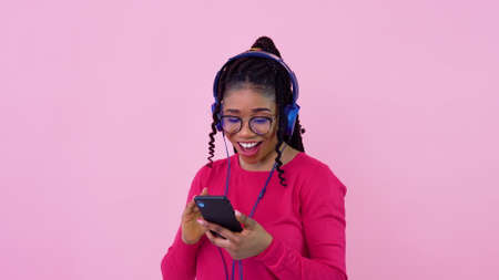 African american girl is listening to music from smartphone in photo studio. Teen girl in pink clothes on a solid pink backgroundの写真素材