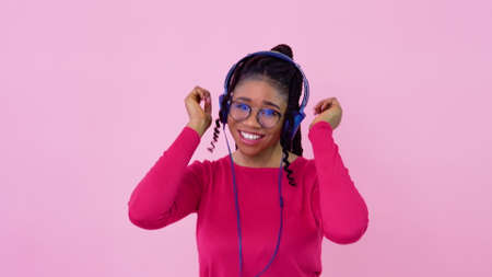 Cute young african american music lover girl is listening to music from smartphone in photo studio. Teen girl in pink clothes dances on a solid pink backgroundの写真素材