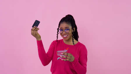 Cute cheerful young african american girl in pink clothes holds the key to the car and puts it in a toy shopping basket. Teen girl housewife beginner standing on a solid pink backgroundの写真素材