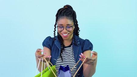 Cute cheerful young african american girl in blue clothes looks in laminated cardboard bags. Teen...