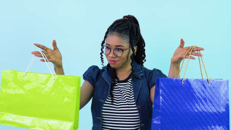 Cheerful young african american girl in blue clothes holding laminated paper bags with pens. Teen girl standing on a solid light blue backgroundの写真素材