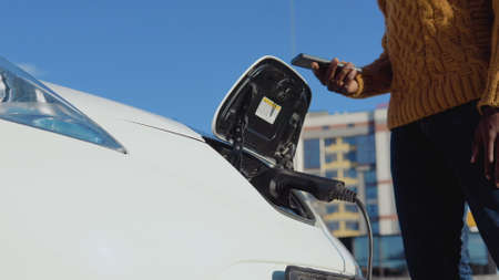 African american male driver connects an electric car to the power system to charge the car battery and controls the charging process via his smartphoneの写真素材