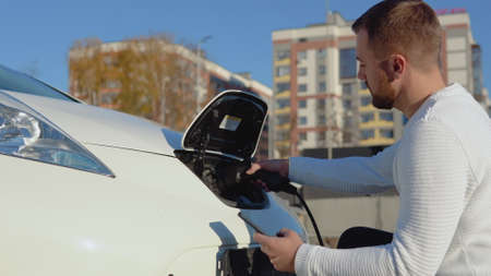A fair-skinned male driver connects an electric car to the power system to charge the car battery and controls the charging process via his smartphoneの写真素材