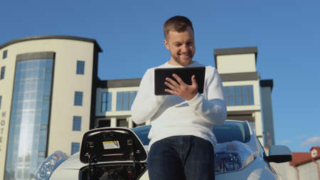 A light-skinned male driver stands with a tablet in his hands near an electric car while it is chargingの写真素材