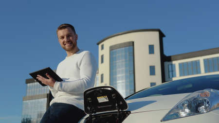 A male driver stands with a tablet in his hands near an electric car while it is chargingの写真素材